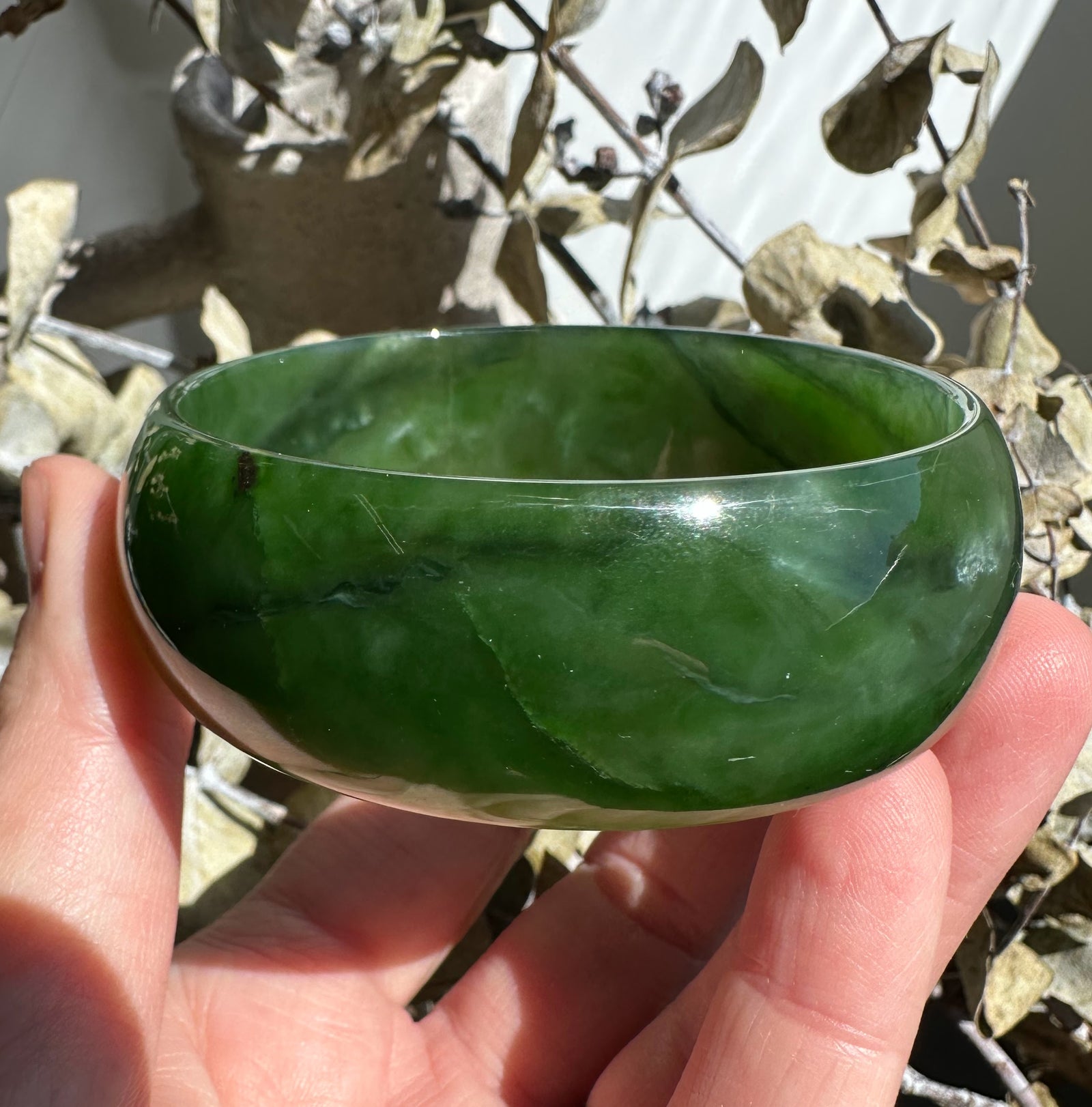 Close up of a glossy green jade bangle held between fingers, showing deep marbling and color variations.