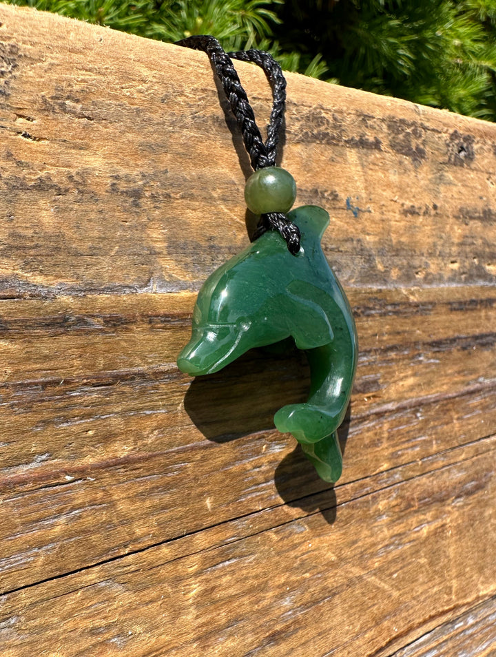 Green jade dolphin pendant hanging from a braided black cord with a bead on a weathered wooden surface