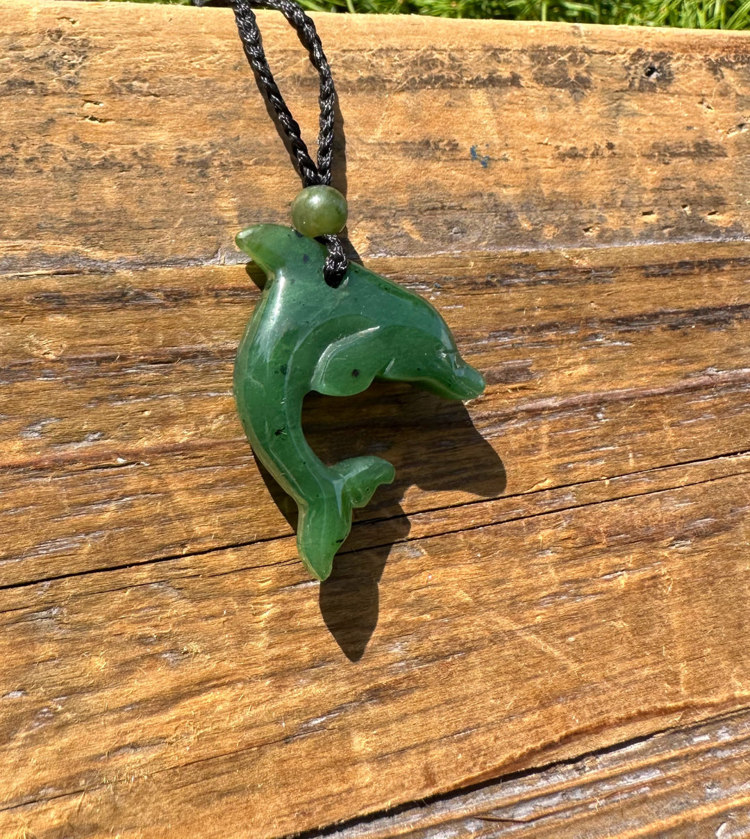Green jade dolphin pendant attached to a black braided cord with a green bead shown from a side angle on wood.