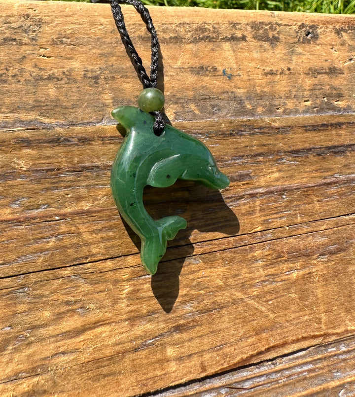 Green jade dolphin pendant attached to a black braided cord with a green bead shown from a side angle on wood.