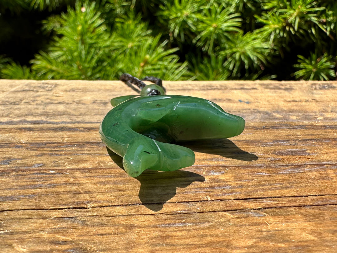 Green jade dolphin pendant with a black cord and green beads resting on a weathered wooden surface.