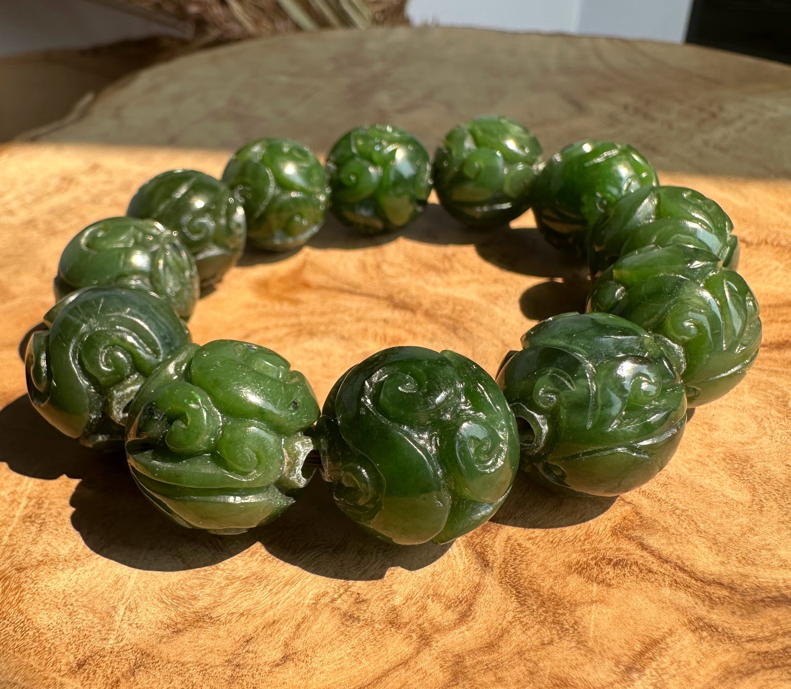 A beaded bracelet made of carved green jade-like round beads with swirling engravings, displayed on a textured light brown wooden surface.