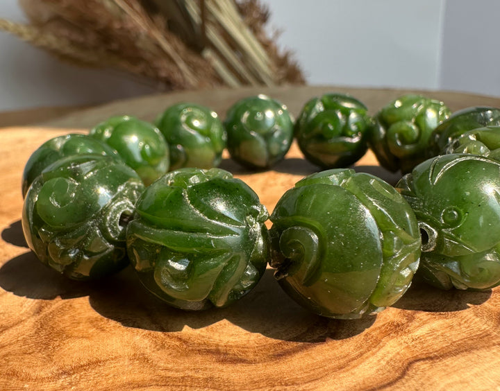 A beaded bracelet made of glossy carved green jade beads, displayed on a wooden surface with dried grass in the background.
