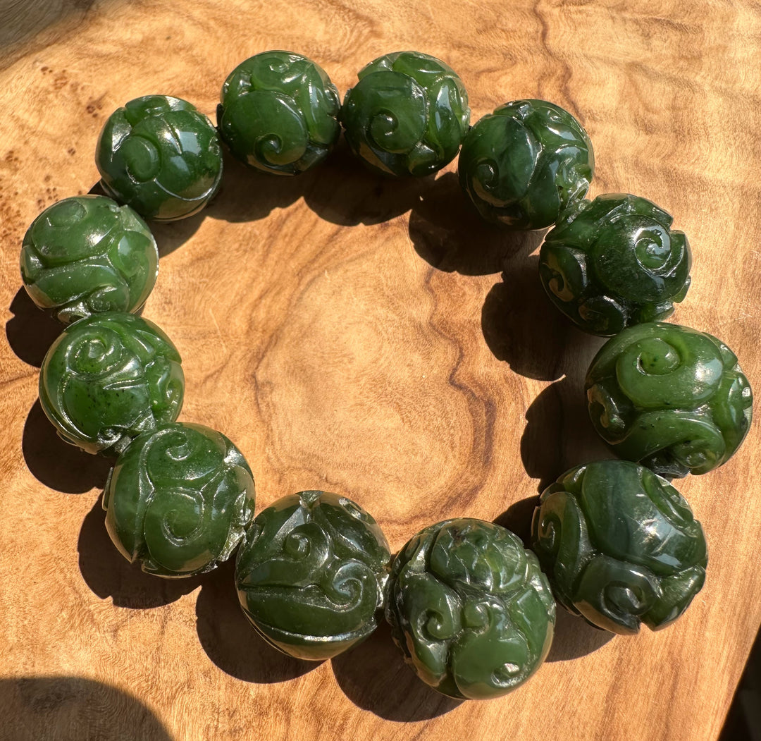 A circular beaded bracelet made of carved dark green jade round beads with swirling floral or cloud motifs, photographed on a warm wood surface.
