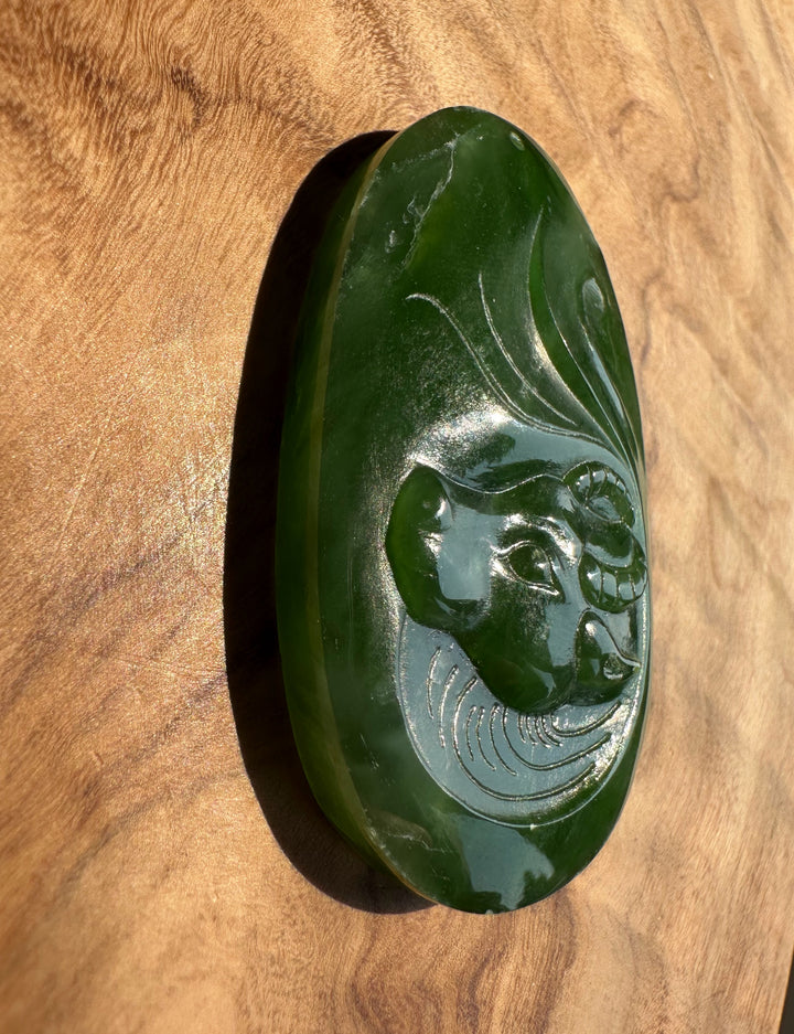 A polished dark green jade pendant featuring a carved buffalo face, displayed on a warm wood grain background.