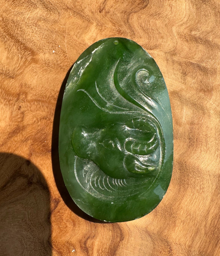 A green jade carved pendant depicting a stylized buffalo, displayed on a textured wooden surface.