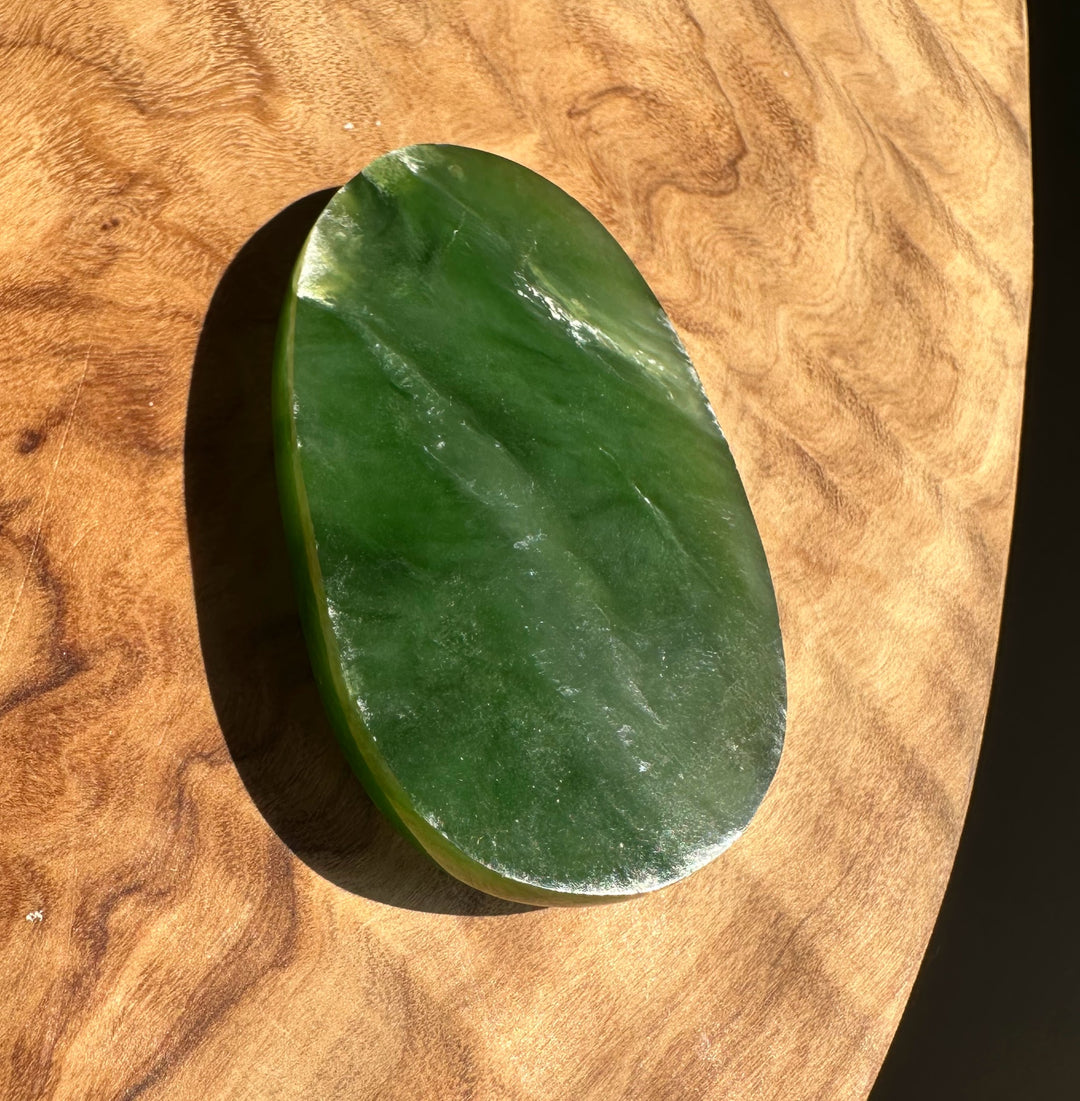 A polished green stone or jade-like oval object with a smooth glossy surface, photographed on a curved wooden surface with direct sunlight casting a shadow.