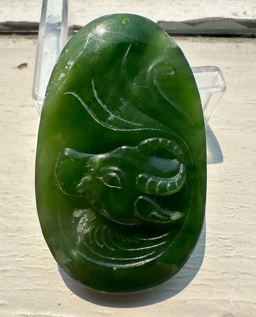 A green carved stone pendant or plaque featuring a stylized animal head with flowing lines, displayed on a clear stand against a light off-white surface.