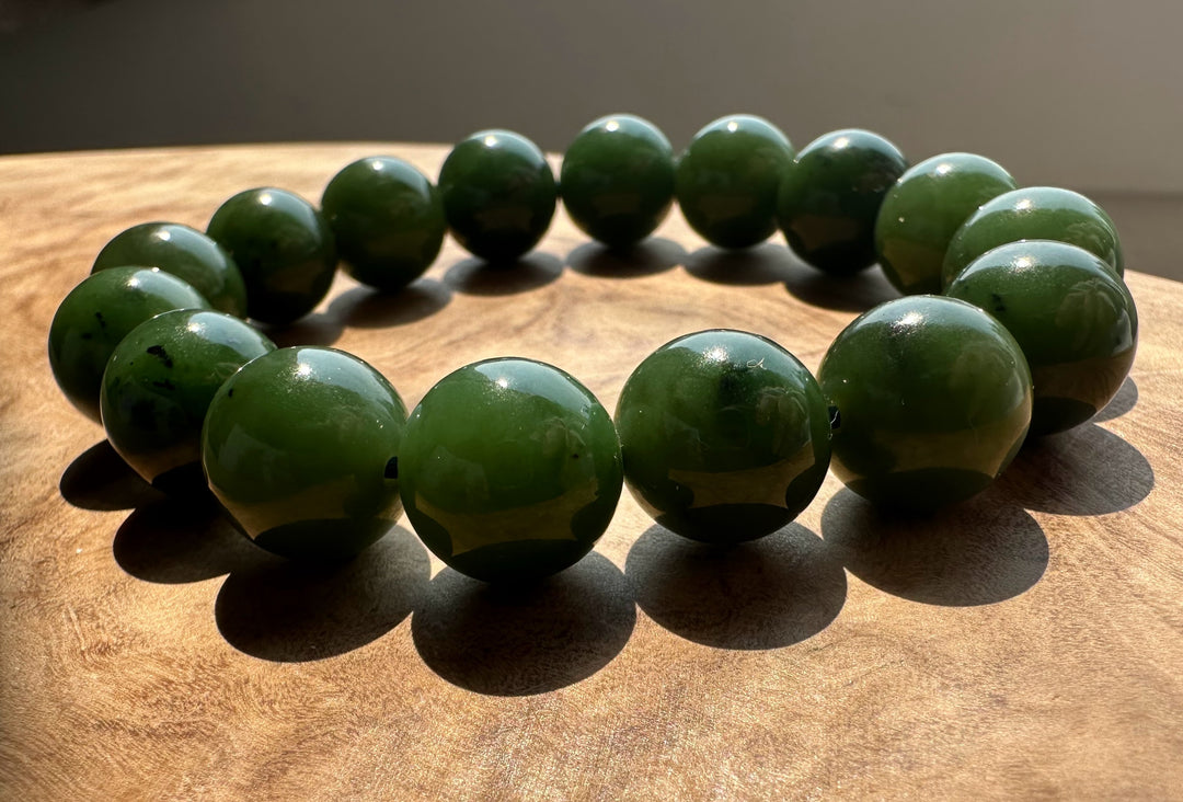 A beaded bracelet made of polished dark green jade round beads, arranged in a circle on a wooden surface with natural sunlight casting shadows.