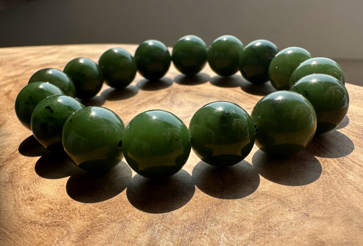 A beaded bracelet made of polished dark green jade round beads, arranged in a circle on a wooden surface with natural sunlight casting shadows.