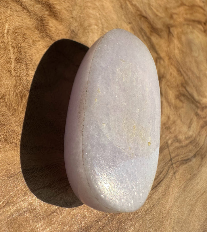 A smooth, polished oval stone with a pale lavender-purple hue and subtle metallic sheen, resting on a textured brown wooden surface.