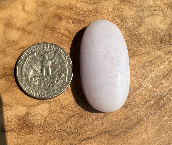 A smooth, polished light purple jadeite oval stone placed next to a US quarter coin for scale, set on a textured wooden surface.
