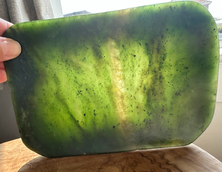 A hand holding a large, polished green jade with rounded corners and a natural marbled texture, set against a background of a window and wooden table.