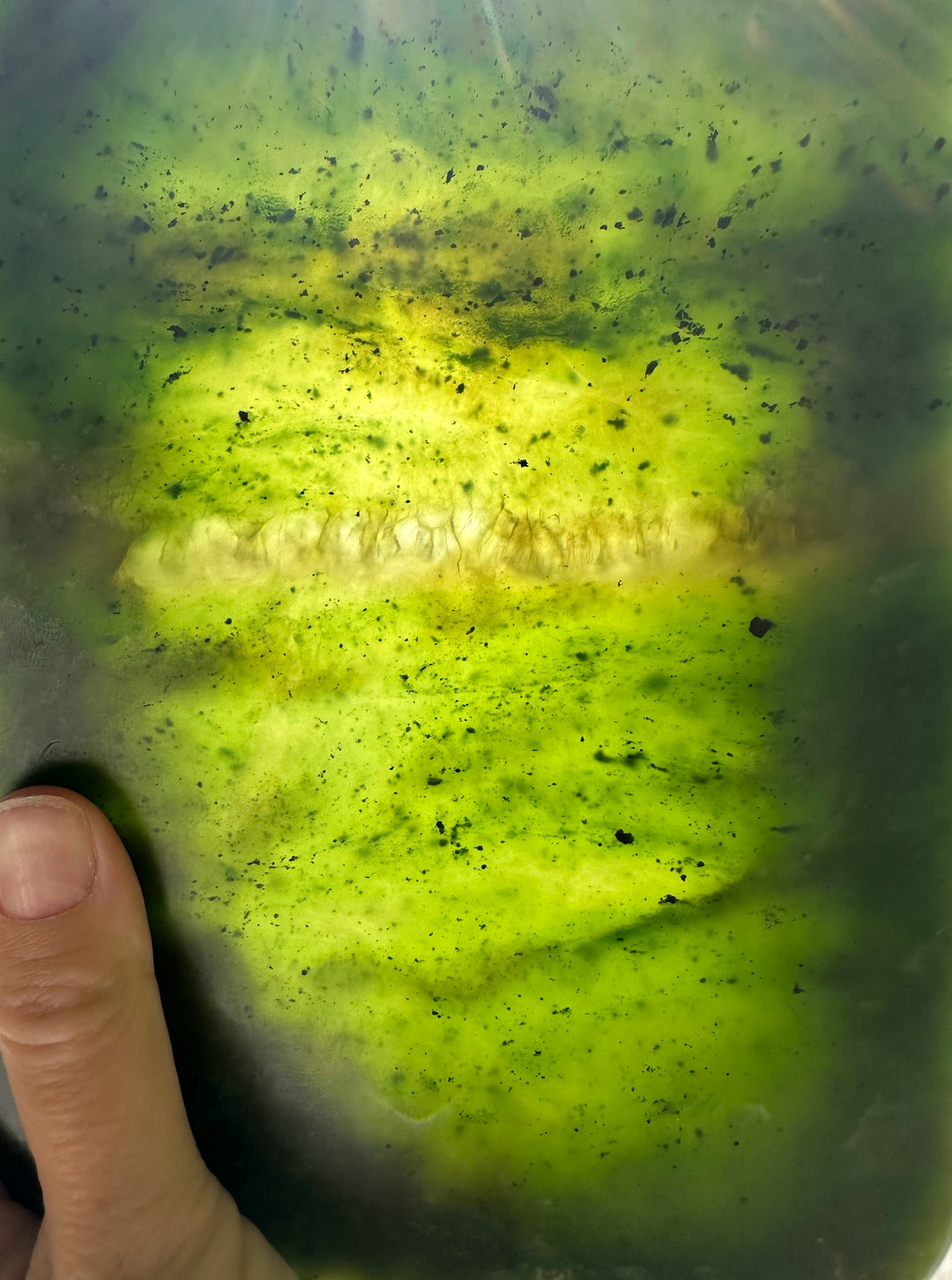 A close-up of a green jade held by a hand, showing a long, pale yellow-white stripe running horizontally through the center with dark speckles.