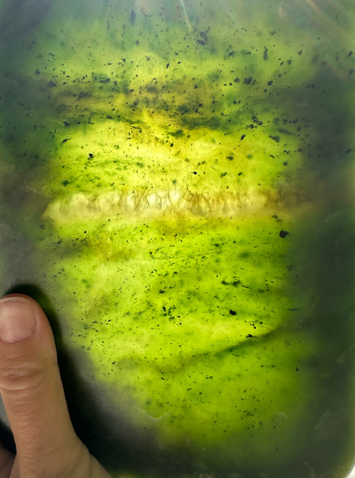 A close-up of a green jade held by a hand, showing a long, pale yellow-white stripe running horizontally through the center with dark speckles.