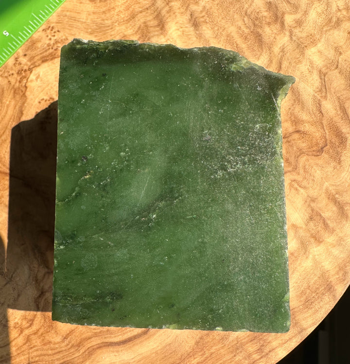 A rectangular green jade with rough, uneven edges, photographed on a curved wooden surface with a small green ruler visible in the top-left corner.
