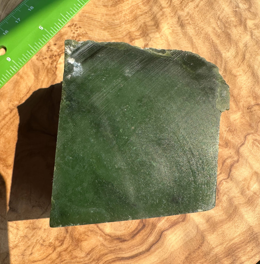 A polished green stone with natural irregular edges, photographed on a wood-grain surface with a green ruler partially visible in the top left for scale.