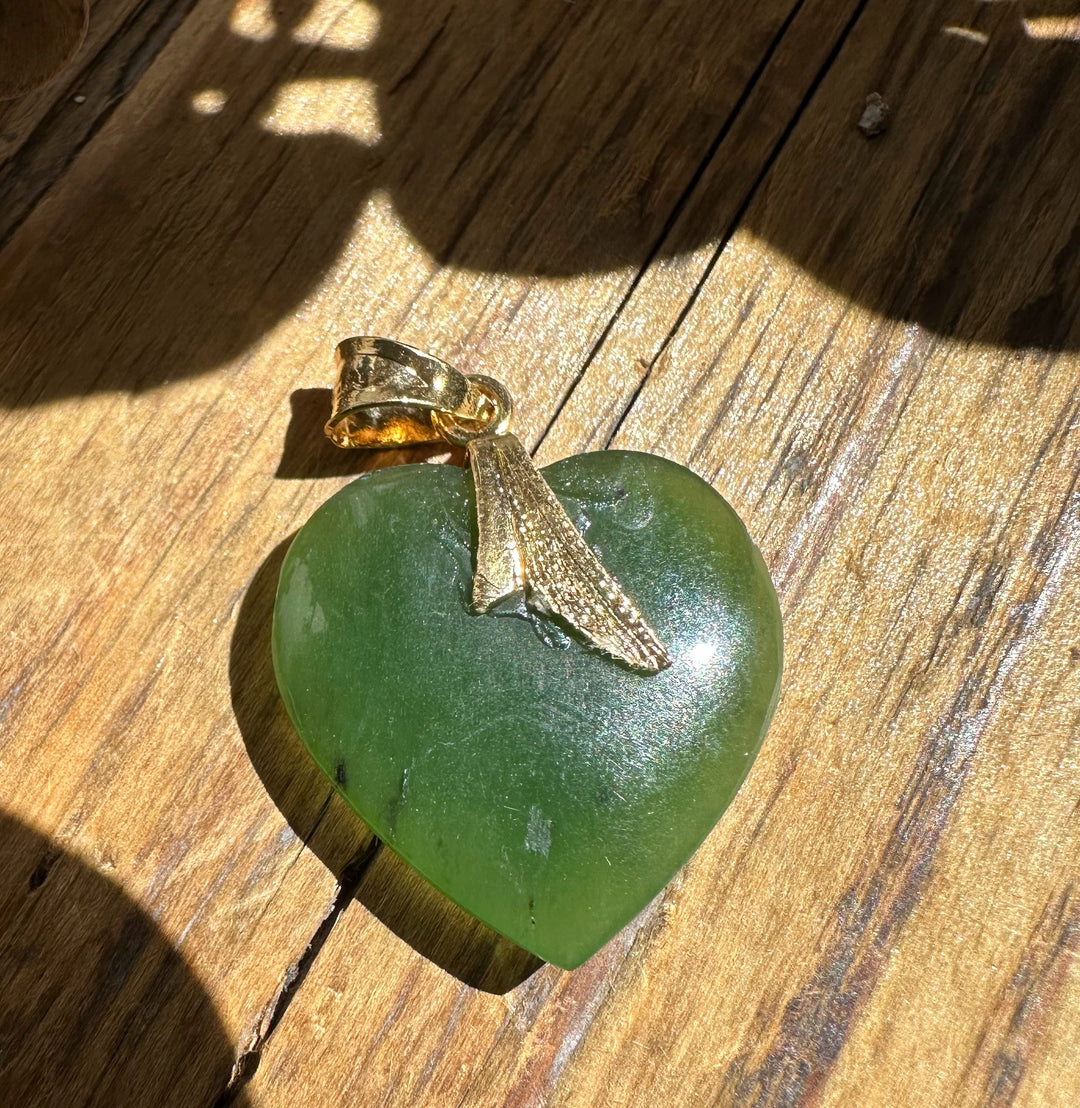 Green jade heart pendant with a gold bail and leaflike cap on wood.