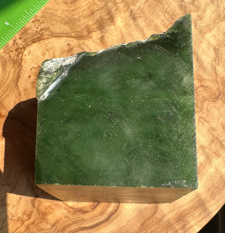 A green jade stone block with a rough, natural uncut edge, resting on a burl wood surface with a green ruler visible in the top-left corner for scale.