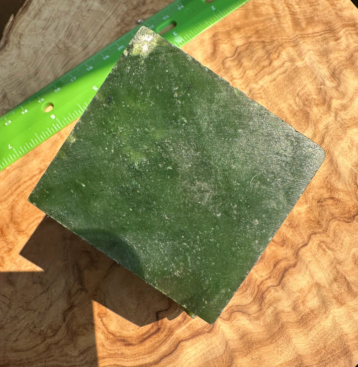 A square green jade tile or slab shown on a wavy wood surface with a green ruler for scale.