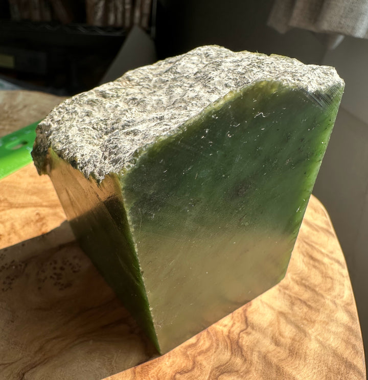 A rectangular green jade with a rough, textured gray top, resting on a wooden surface in warm natural light.
