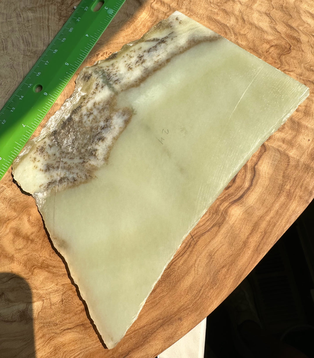 A raw, irregularly shaped slab of pale green jade with a mottled brown and cream pattern along one edge, displayed on a wooden surface next to a green ruler.