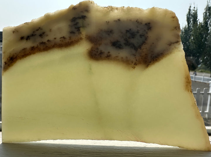 A translucent pale yellow jade slab with dark brown to black mottled areas near the top, photographed outdoors against a blurred background of trees and railing