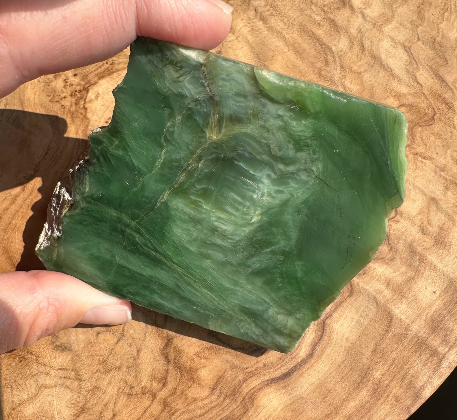A hand holding a raw green jade with natural marbled patterns, set against a polished wood background.
