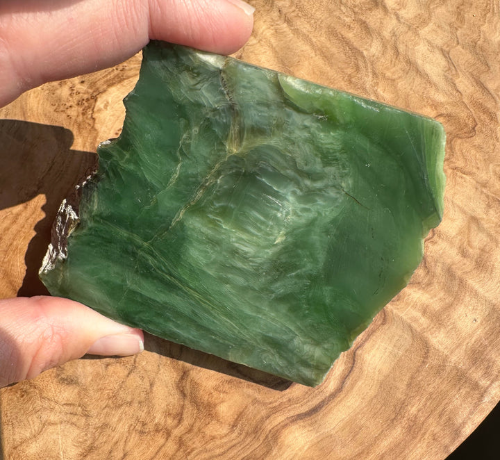 A hand holding a raw green jade with natural marbled patterns, set against a polished wood background.