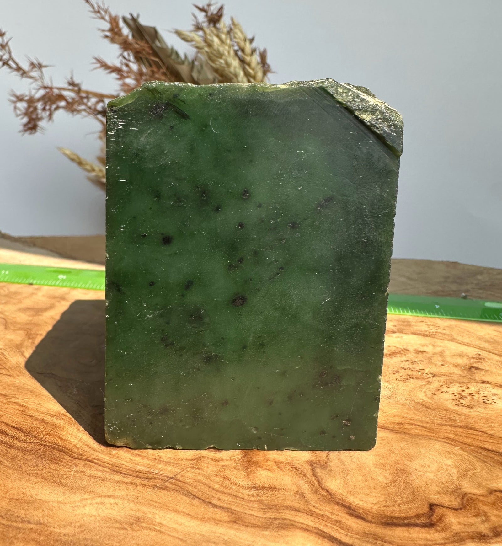 A rectangular green polished stone or jade-like block with a rough chipped edge, standing upright on a wooden surface with a green measuring tape visible behind it.