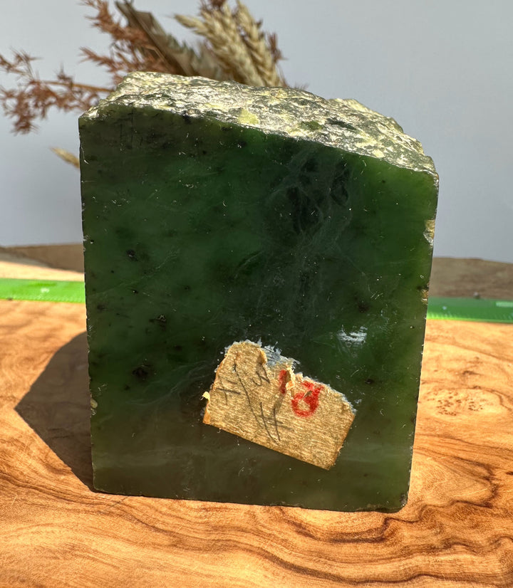 A rough, polished green stone block or decorative slab with natural marbling and dark inclusions, standing upright on a wooden surface. A small beige paper tag with a red &