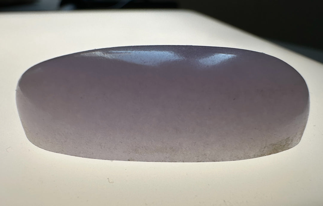 A smooth, polished oval-shaped translucent stone object in a pale lavender-purple hue, resting on a light-colored surface.