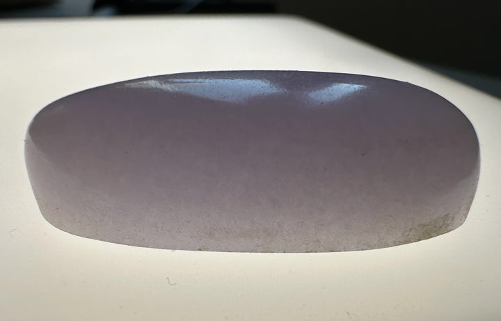 A smooth, polished oval-shaped translucent stone object in a pale lavender-purple hue, resting on a light-colored surface.