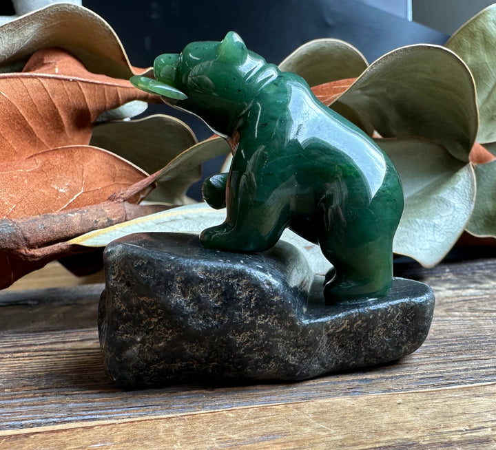 2 inch jade bear sculpture perched on a dark granite base with dried leaves in the background.