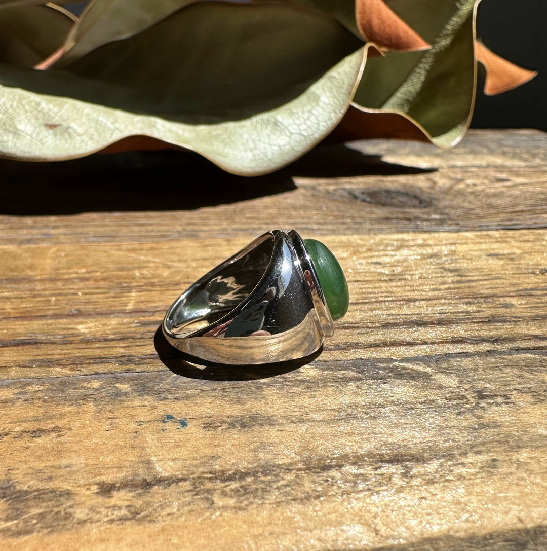 Bold silver ring with a green jade cabochon on a wooden surface bathed in sunlight.