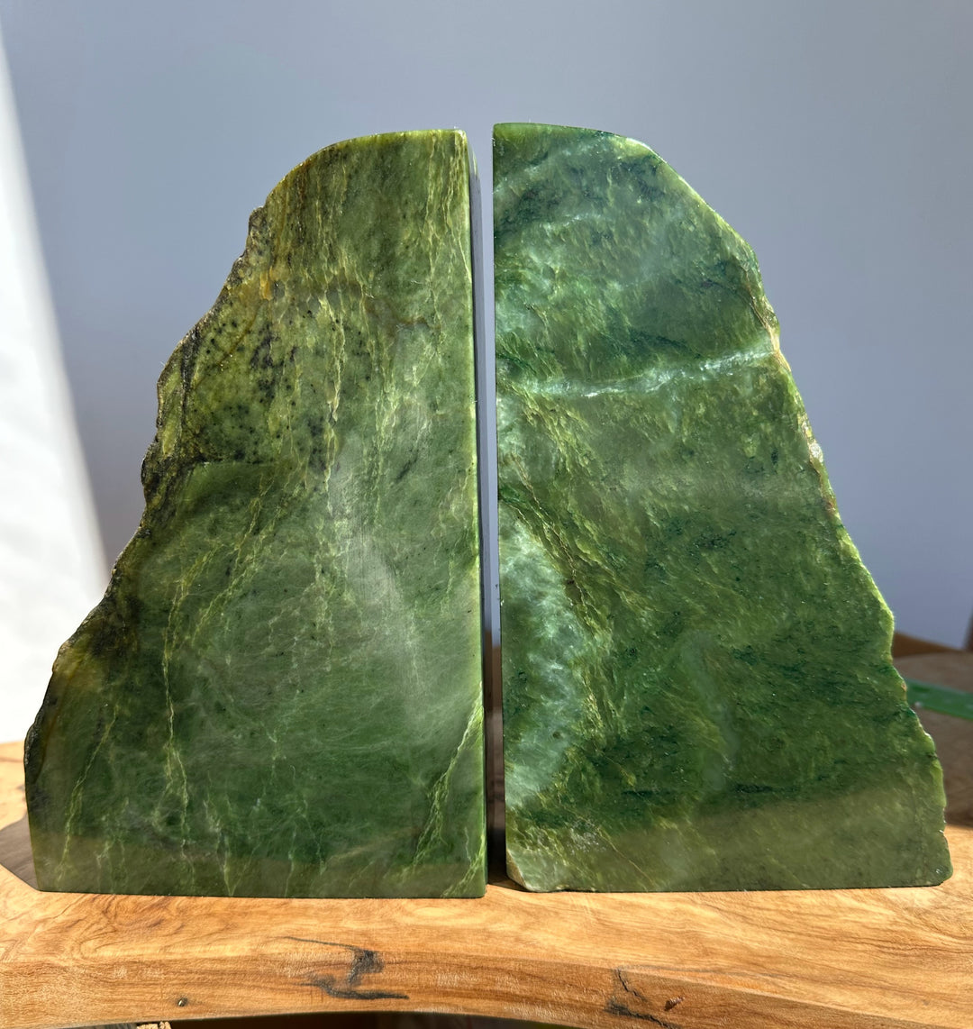 A pair of polished green jade bookends shaped like rough natural slices, displayed on a wooden surface against a soft gray background.