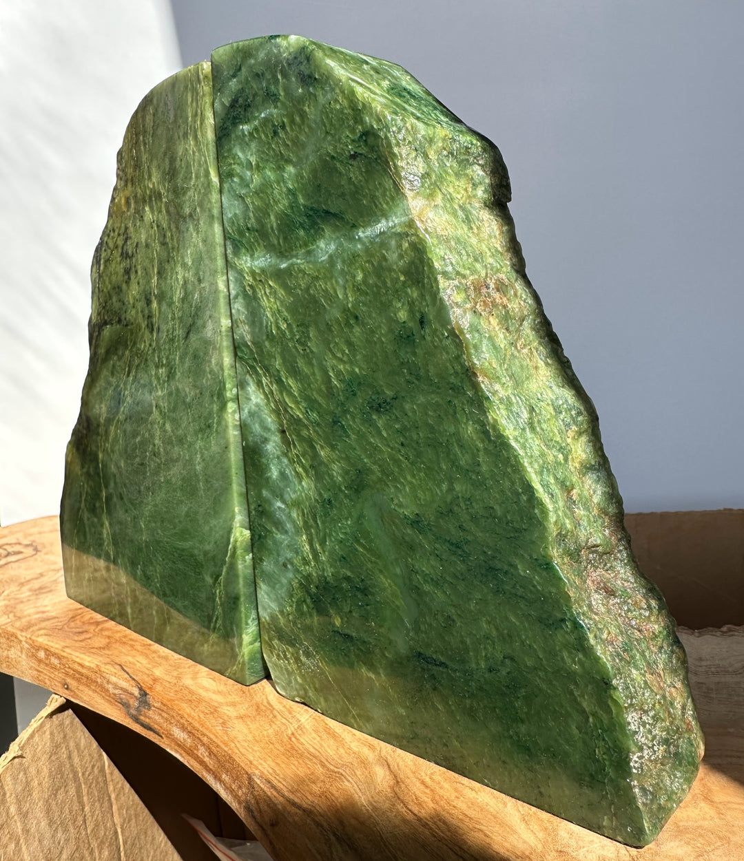A polished green jade bookends with natural marbling and rough edges, displayed on a wooden surface against a plain light gray background.