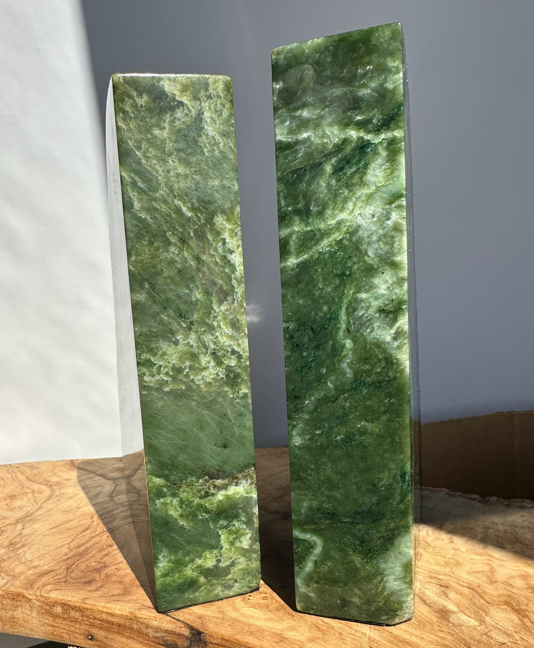 Two polished green stone rectangular blocks with natural marbled texture, standing upright on a wooden surface against a gray wall.