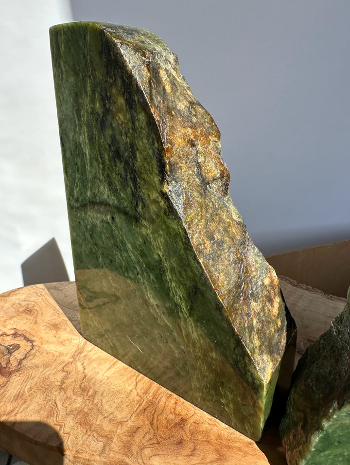 A polished natural green jade bookend with rough, uneven edges and yellow-brown mineral variation, displayed upright on a wooden surface against a plain light gray background.