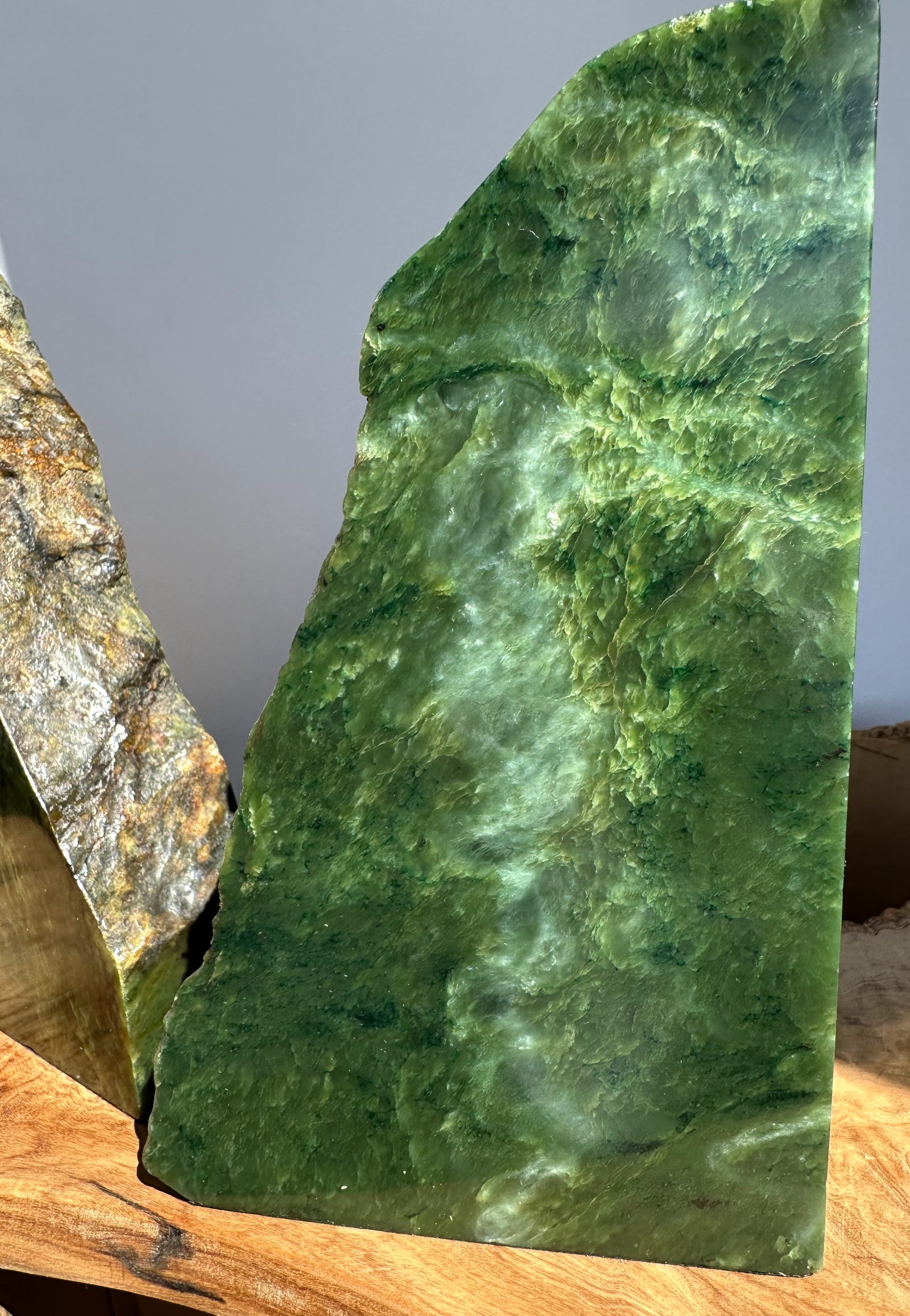A polished green jade stone slab with natural marbling, standing upright on a wooden surface next to an unpolished brown and gold stone.