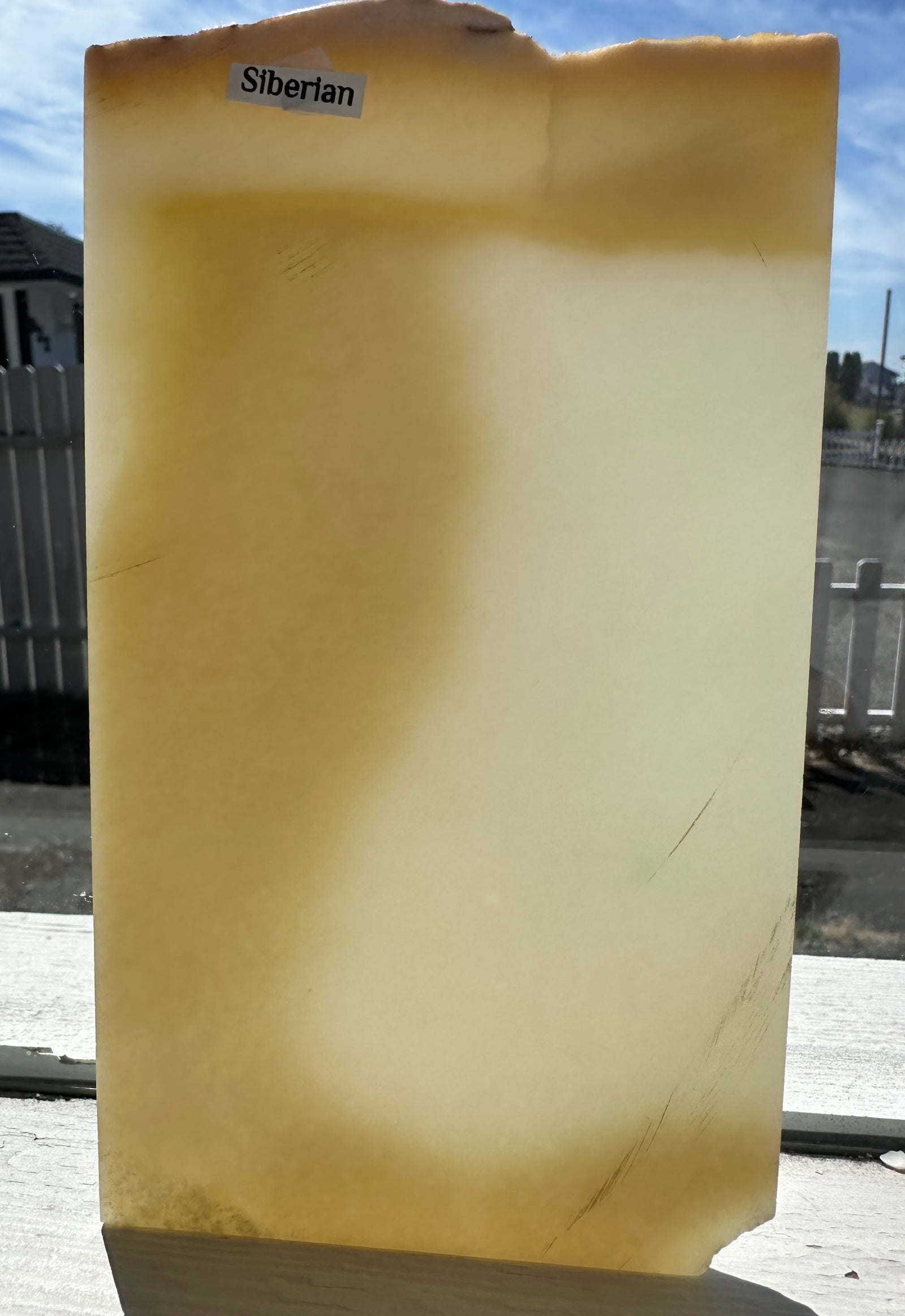 A Siberian jade slab with a warm yellow and cream coloration, standing upright outdoors on a windowsill with a label sticker at the top.