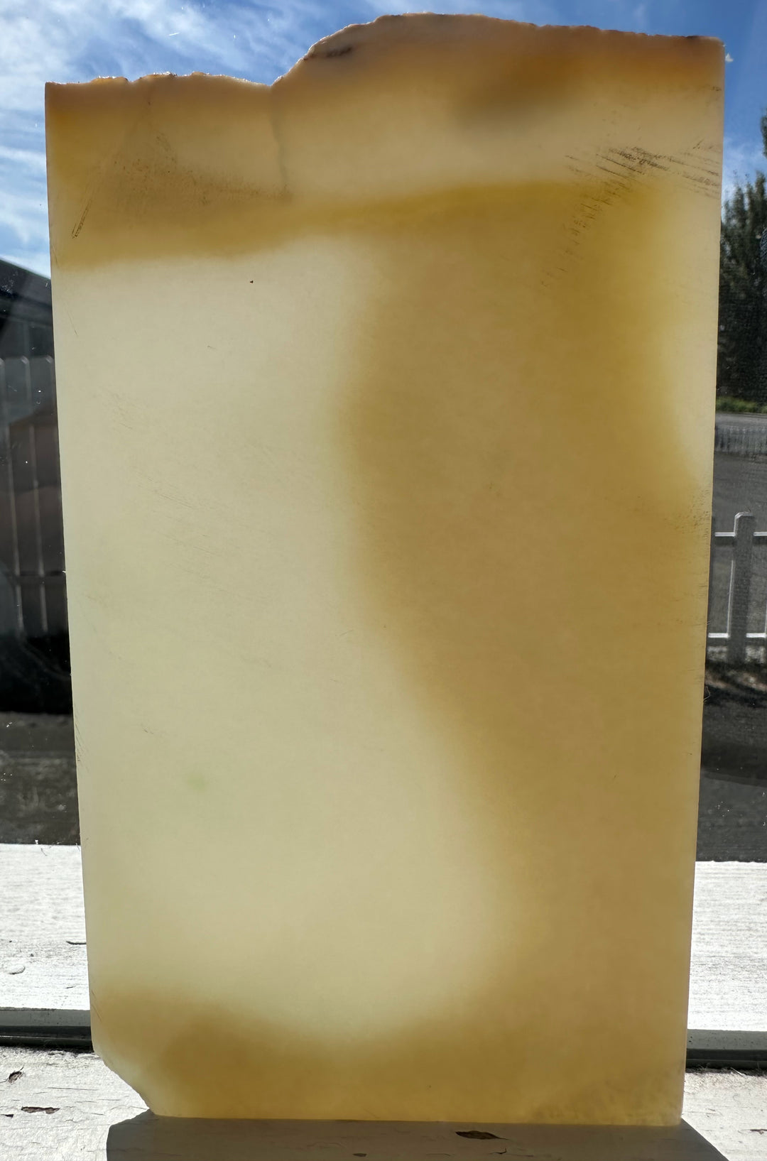 A large, rectangular translucent stone slab with a warm yellowish-beige color and natural rough edges, photographed outdoors against a blue sky and white fence background.