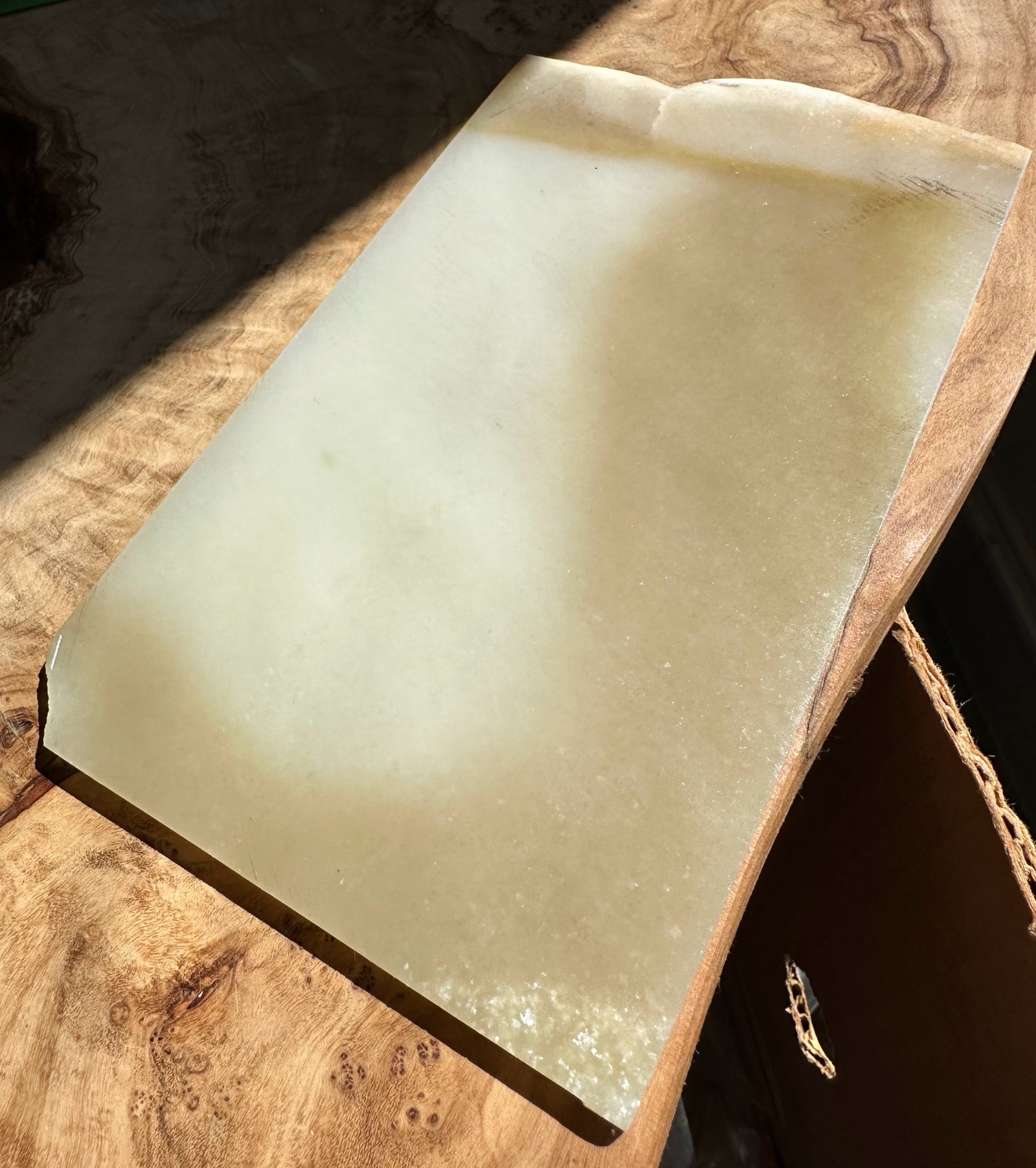 A rectangular translucent jade slab with a pale greenish-gray hue, resting on a rustic wooden surface under direct sunlight.