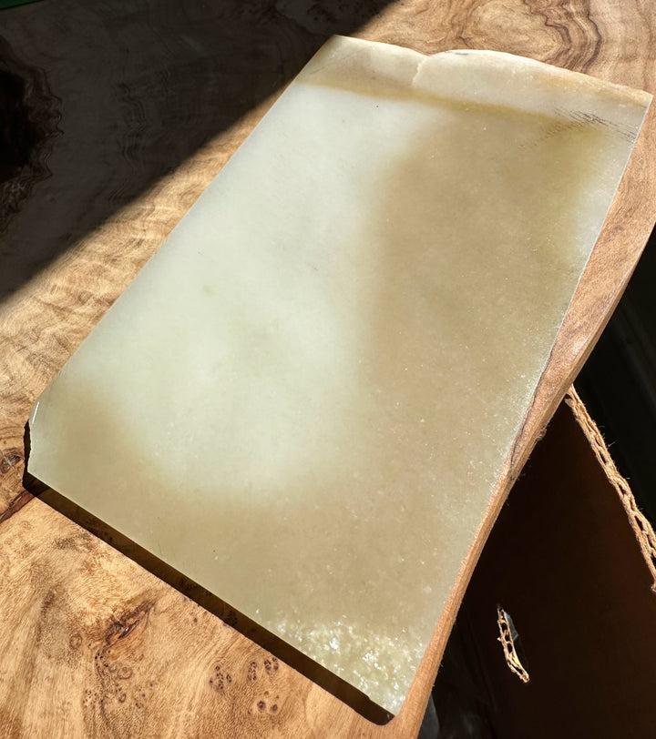 A rectangular translucent jade slab with a pale greenish-gray hue, resting on a rustic wooden surface under direct sunlight.