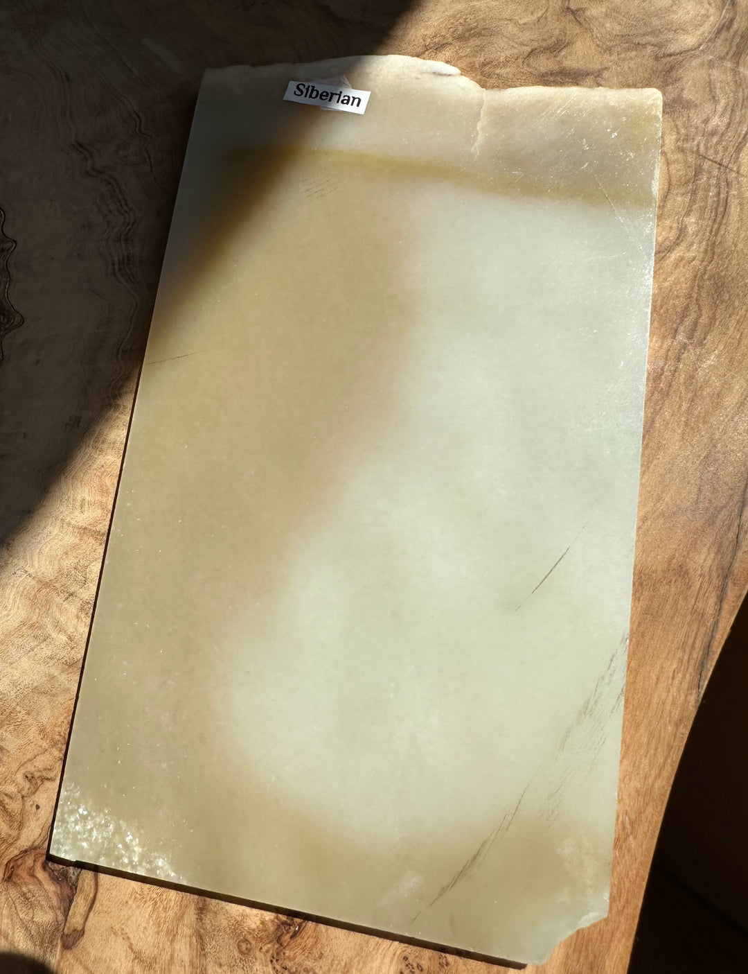 A slab of Siberian jade with a smooth, matte finish, resting on a rustic wooden surface. The stone has a warm, creamy-beige hue with subtle natural variations and a small label at the top reading &