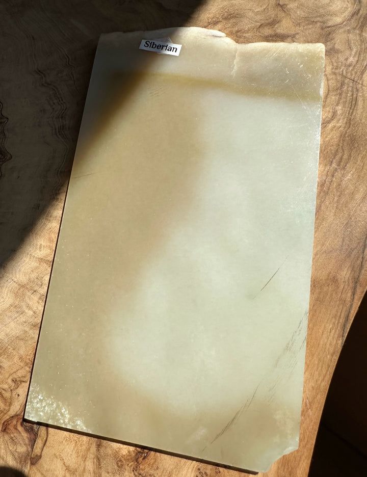 A slab of Siberian jade with a smooth, matte finish, resting on a rustic wooden surface. The stone has a warm, creamy-beige hue with subtle natural variations and a small label at the top reading &