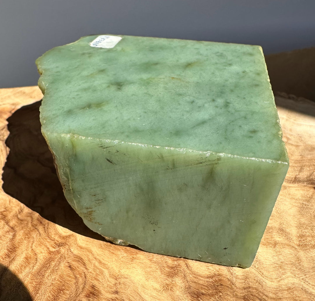 A rough, rectangular green stone block with natural marbling and a small white sticker label, resting on a textured wooden surface.