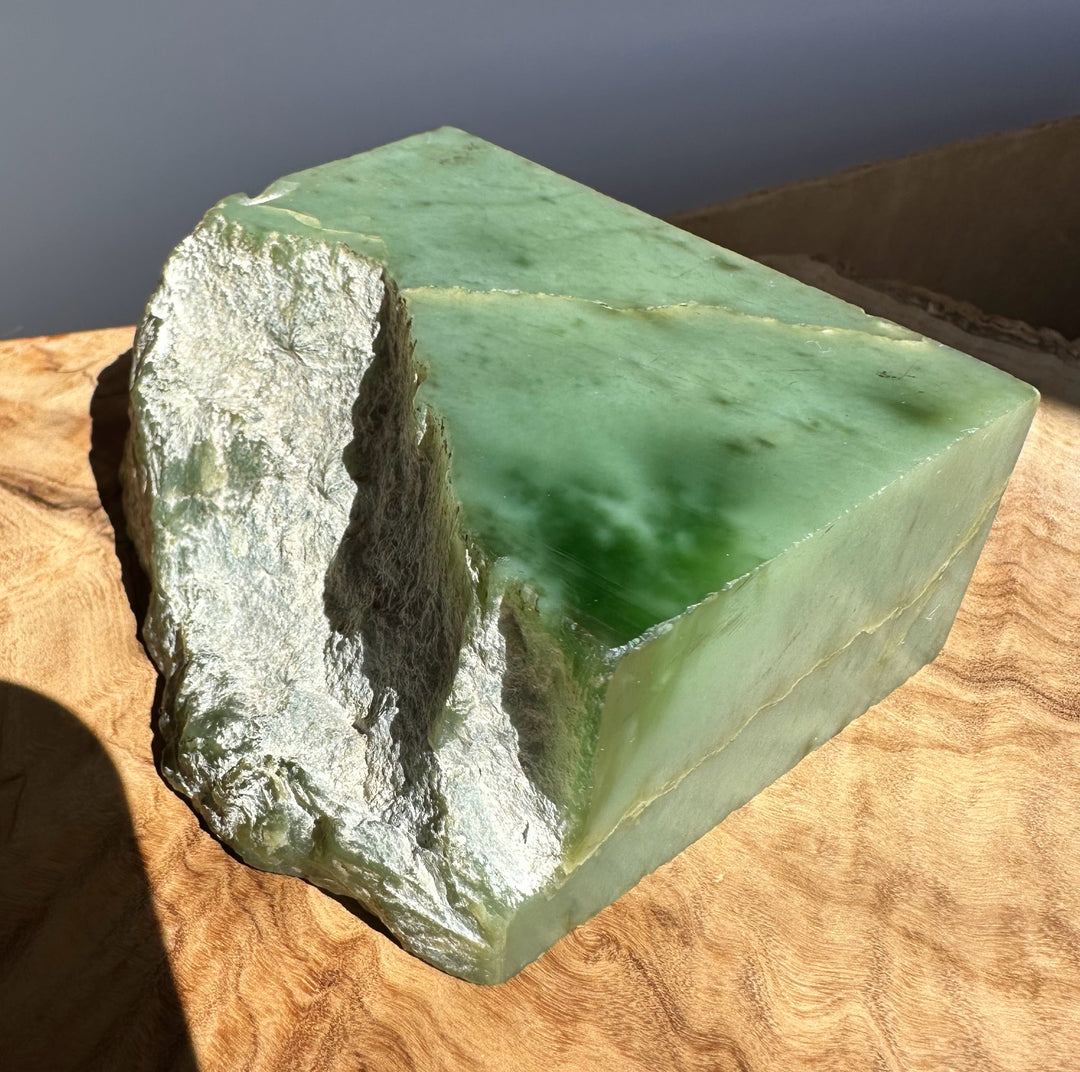 A green jade block with rough, uneven natural edges, resting on a wavy wood surface under direct sunlight.