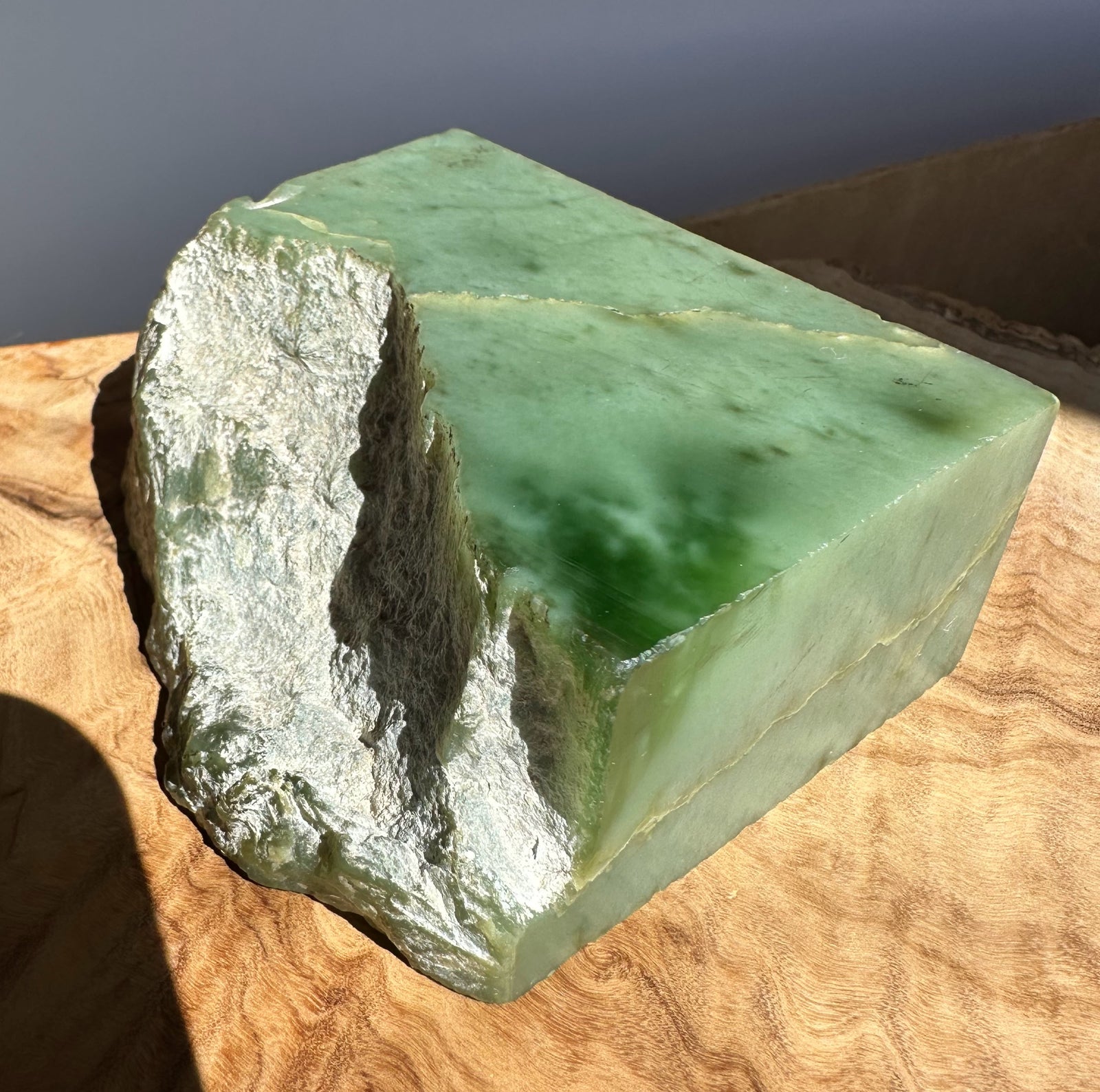 A green jade block with rough, uneven natural edges, resting on a wavy wood surface under direct sunlight.