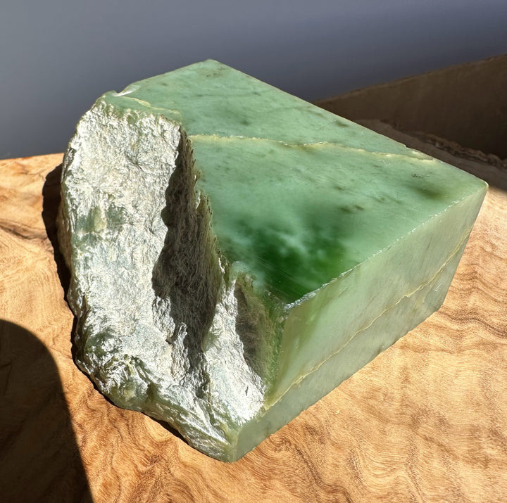 A green jade block with rough, uneven natural edges, resting on a wavy wood surface under direct sunlight.